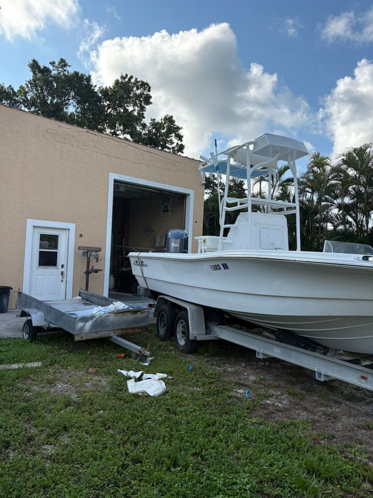 White fishing boat on trailer parked next to brick building with white door and palm trees