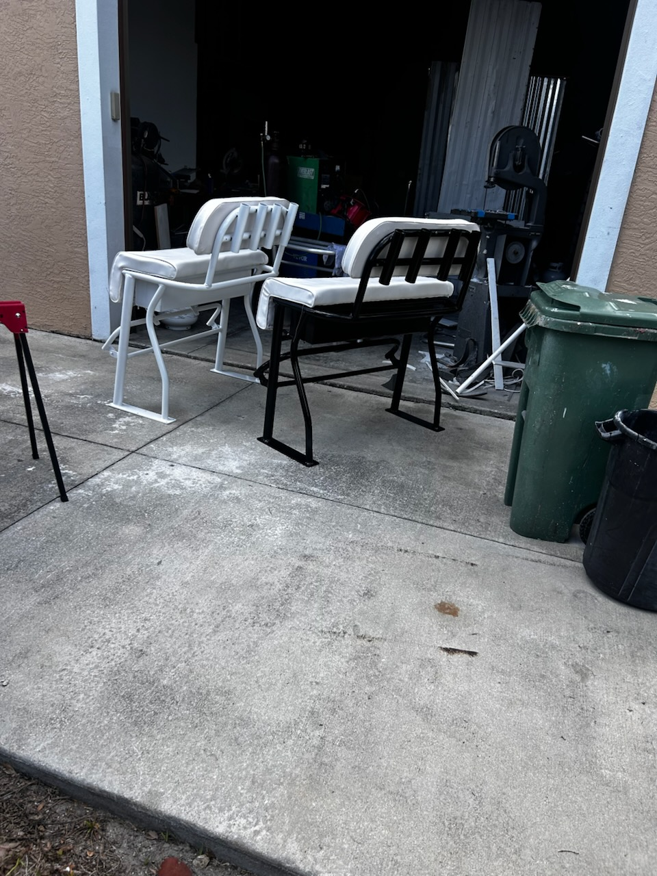 Two outdoor patio chairs with cushions positioned on concrete driveway near garage entrance with trash bins